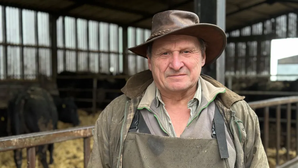 A man in brown farming overalls and a brown wax coat with a brown hat standing in front of a barn of cows.
