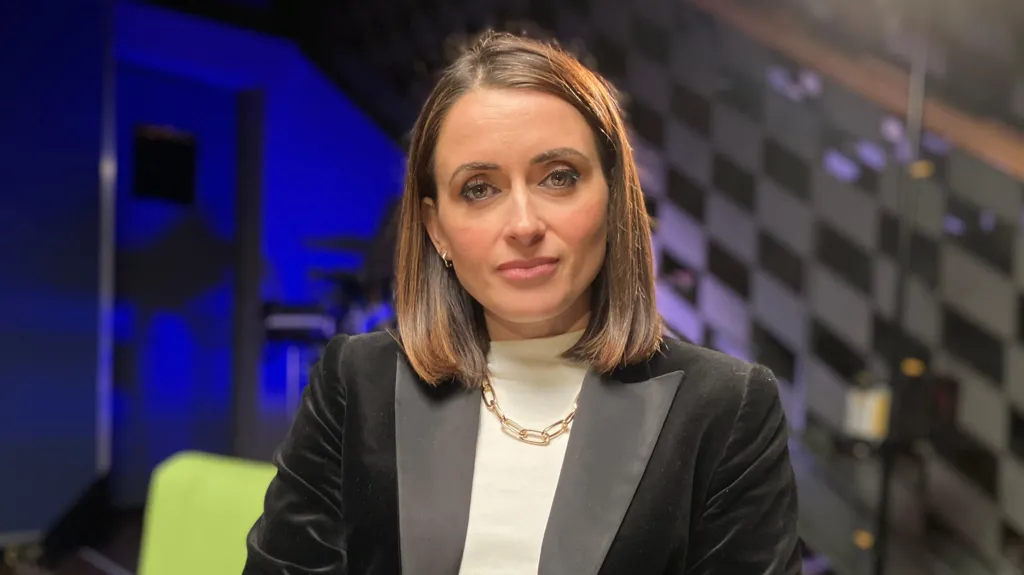 Anne McAlpine, in a white top, gold necklace and black velvet jacket looks dead-on into the camera. She is in a well-lit TV studio with a green chair behind her and a glass wall with a checkered pattern etched into it.