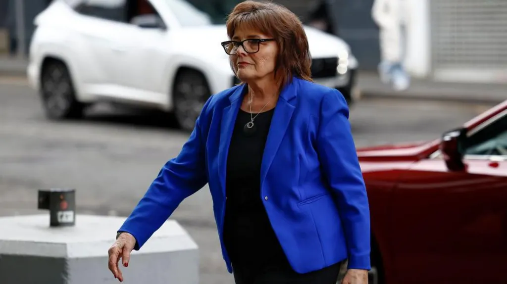 Jeane Freeman, in electric blue jacket and black blouse, is walking purposefully from a car, right to left. She is wearing glasses and looks very serious. A white car can be seen in the background.
