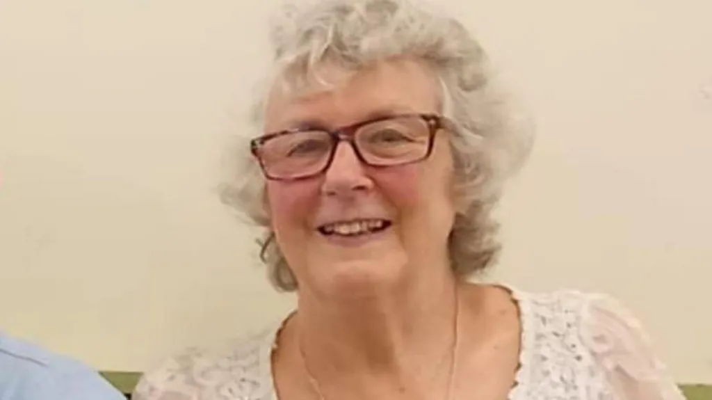 A woman in a white scoop neck lacework top sits inside on a green cushioned bench against a white wall. She is smiling. She is wearing brown framed glasses and her white hair is curly and short.