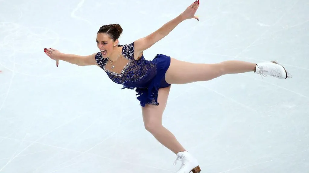 Jenna McCorkell has her brown hair up bun. Her arms are outstretched and she is skating on one skate. She is wearing a dark blue ice-dancing dress which is covered in sequins.  