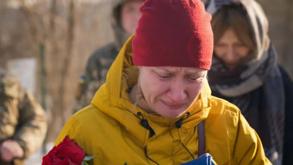Natalia weeps by the grave of her husband in a snow-covered cemetery in Kyiv. She's dressed in a yellow jacket and a red beanie hat, and is holding a red rose. 