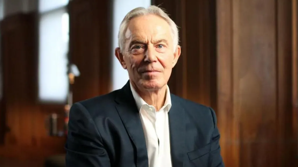 An image of former UK prime minister Tony Blair looking at the camera while dressed in a black suit and white shirt in a room with wooden walls.