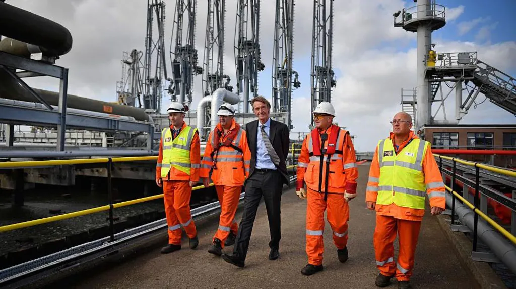 A man in a dark grey suits walks outside on an industrial site with four men in orange high-vis clothing and hard hats. Behind them is port apparatus and machinery on a dock