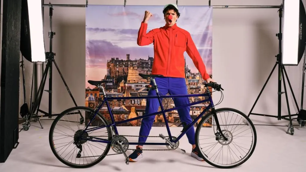 Radio 1 DJ Greg James stood behind a black tandem bike with his left hand on the handlebars and his right arm in the arm with his fish clenched. He is wearing blue trousers and a red jacket and has a red nose on his nose, with a white cap. The photo is taken in a studio setting so there are two big bright photography lights behind him and a canvas style picture of Edinburgh Castle.