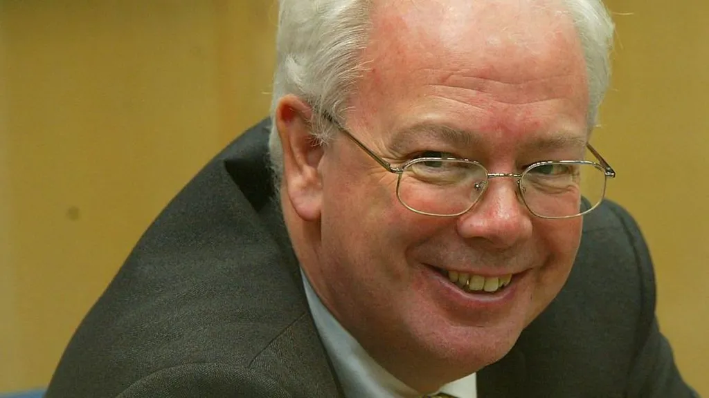 Jim Wallace is leaning forward and smiling broadly at the camera. He has short, grey/white hair and metal-rimmed glasses. He is wearing a dark suit jacket and a white shirt. Only his head and shoulders are showing on the picture
