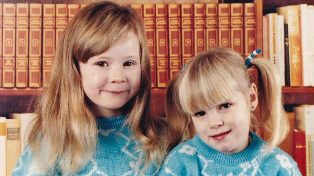 Hayley and Kirsty Wilkinson are two girls aged approximately five and three. Hayley, left, has chest-length blonde hair with a long fringe brushed to the side. Kirsty has blonde hair worn in two bunches. They are wearing identical turquoise jumpers with irregular white lines forming patterns on it. They are both smiling at the camera standing in front of bookshelves.
