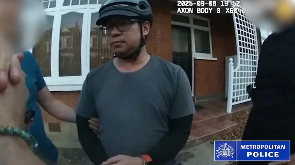 A still from bodycam footage of the police arrest of Vincent Chan. He is outside a house and is weaing a grey T-shirt with a black long-sleeved top underneath. He is wearing glasses and a bicycle helmet. Police are standing either side of him and his arm is being held.