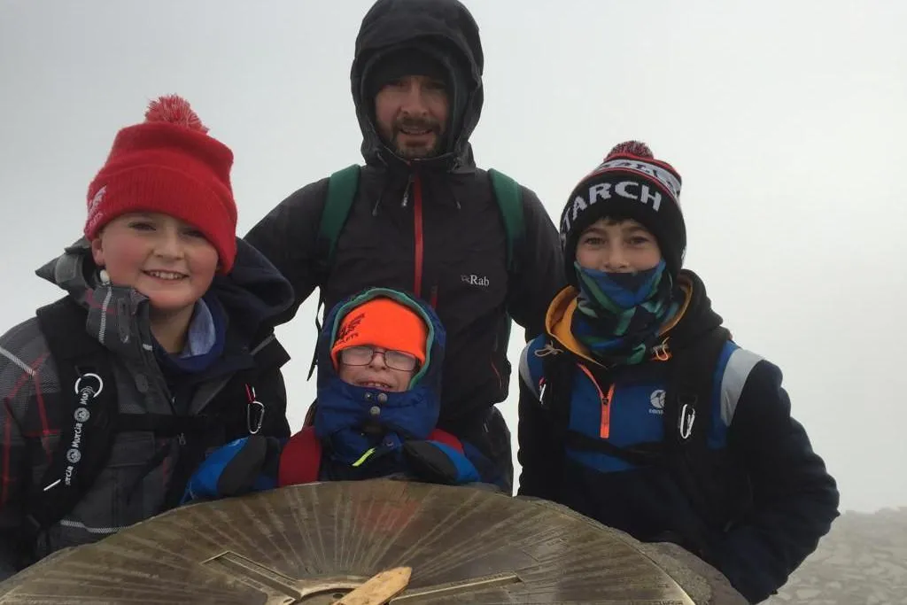 Gerwyn and his three sons are all wearing Scarlets-branded woolly hats, warm coats and rucksacks, and are stood next to the cairn at the peak of Yr Wyddfa. The children are in their early teens and younger. They are all smiling. Visibility is non-existent behind them with a wall of grey cloud.  