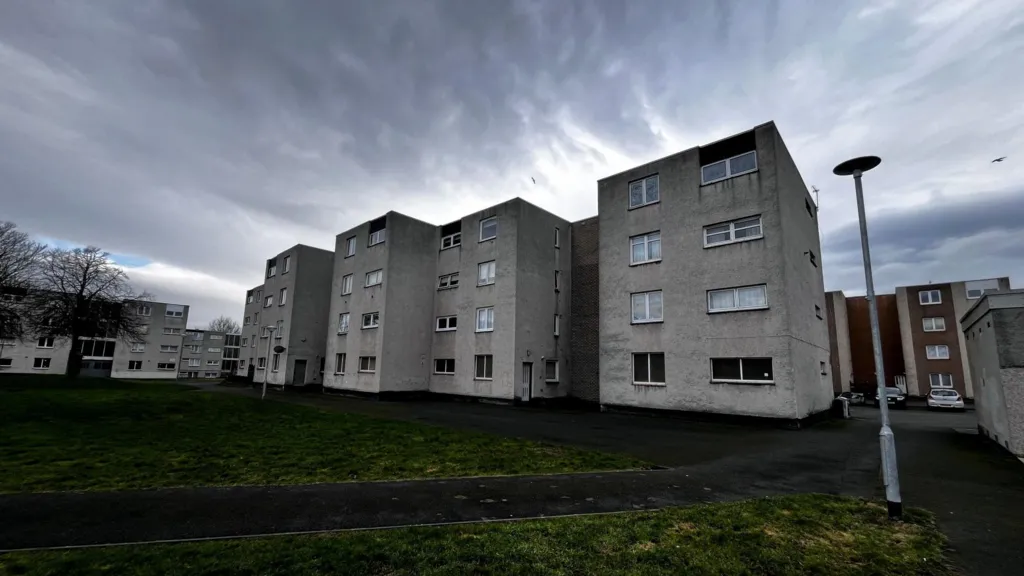 Several blocks of four storey, grey coloured flats with a grassed area to the front.