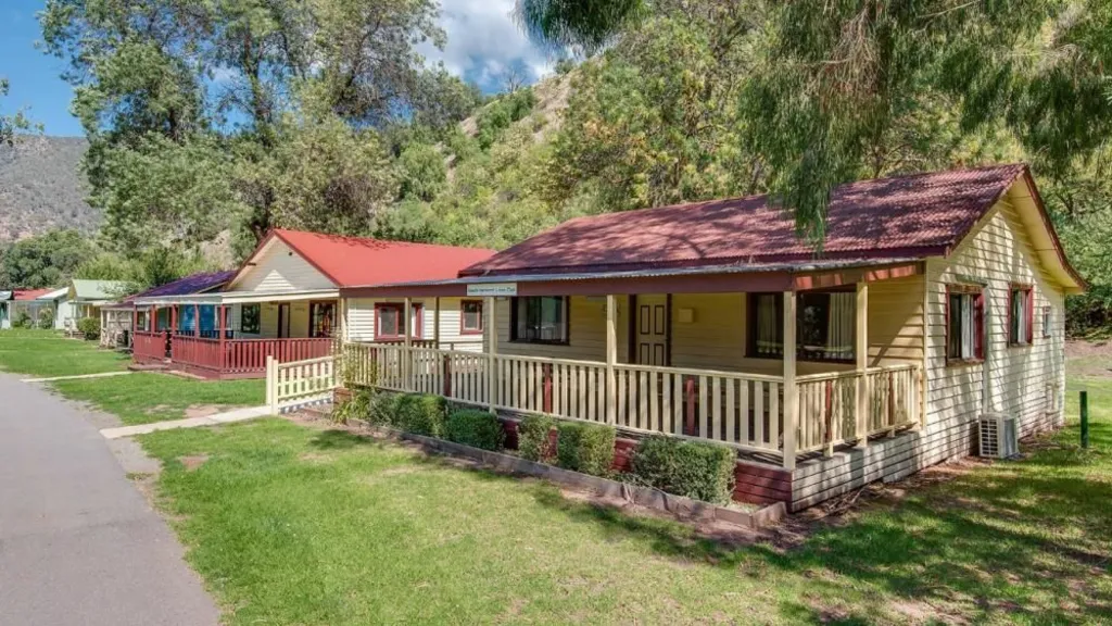 A yellow weatherboard house with a maroon roof surrounded by green grass, in a row of other houses, is pictured in front of a mountain, with trees and shade visible. 