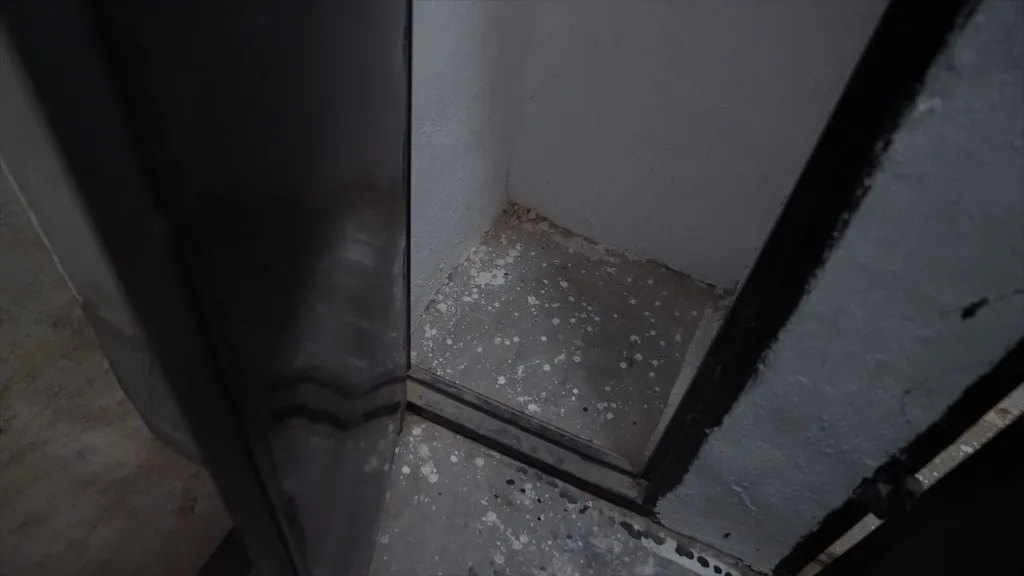 Photo looking down showing the lower part of a cell that is about a metre by a metre in size, with a concrete floor and white-painted walls. Its tall black door is open.
