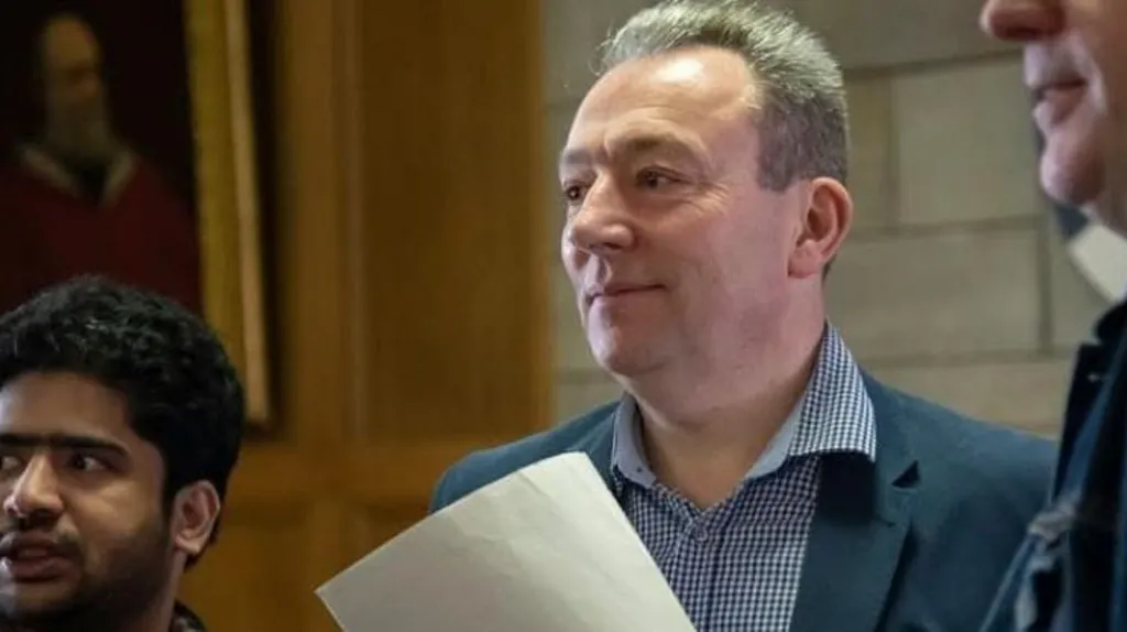 Jim Carruth is a middle-aged man with swept back hair. He is wearing a blue suit, blue and white checked shirt and is standing between two other men who can just be seen on the edge of shot. He is holding a piece of paper.