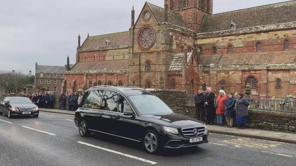 A hearse carrying the coffin of Lord Wallace passes St Magnus Cathedral. People have lined the side of the road