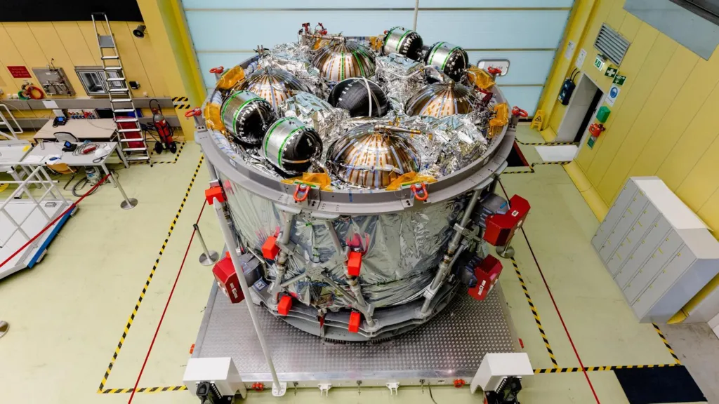 The European Service Module, which is a large cylindrical part of a spacecraft, sits in a cleanroom. On its top surface, several spherical metal tanks are surrounded by metallic insulation. There is shiny silver metallic insulation on its sides too. The module sits on a square platform, and the floor around it is marked with yellow and black safety tape.