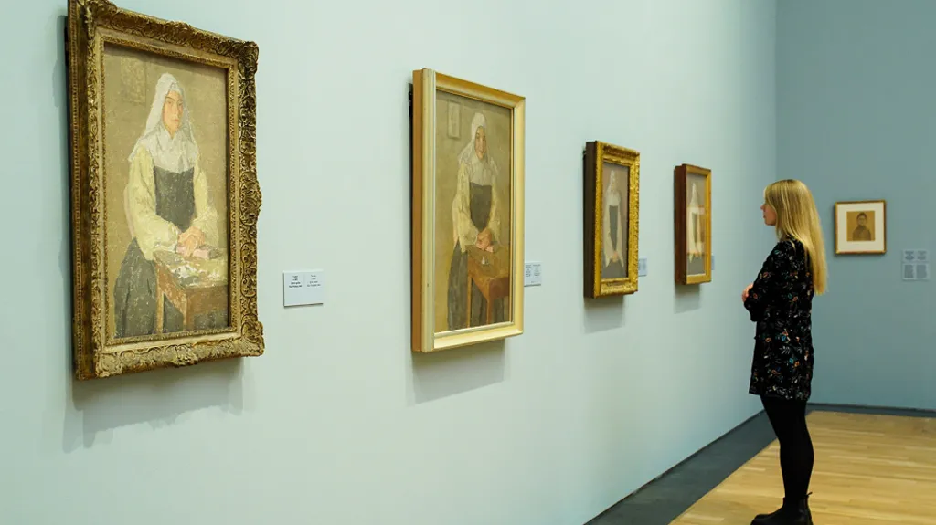 There are a sequence of four similar portraits on the blue wall of a gallery depicting a nun in seated postures, some at a table. Curator Lucy Wood stands in front of the third furthest from the camera looking at the portrait. She is a white woman with long highlighted blonde hair. She wears a black smock dress or shirt with muted flowers as a design over black leggings and black ankle height boots.