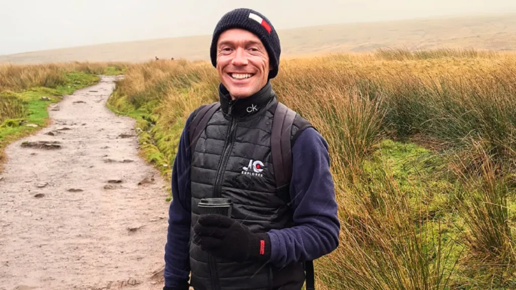 Jay smiling looking at the camera. He is standing on a muddy walking path on a mountain. Jay wears a long sleeve navy top, a navy hat, a black gilet, black gloves and holds a takeaway coffee in his left hand.