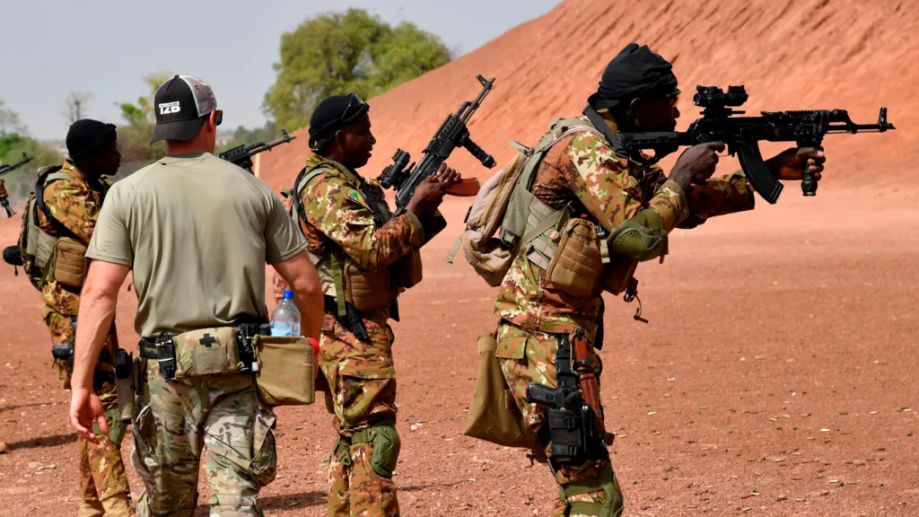 An American military trainer walks past Malian soldiers aiming their guns. They are wearing camouflage and are standing in front of a hill of red earth. The photo was taken in 2018.