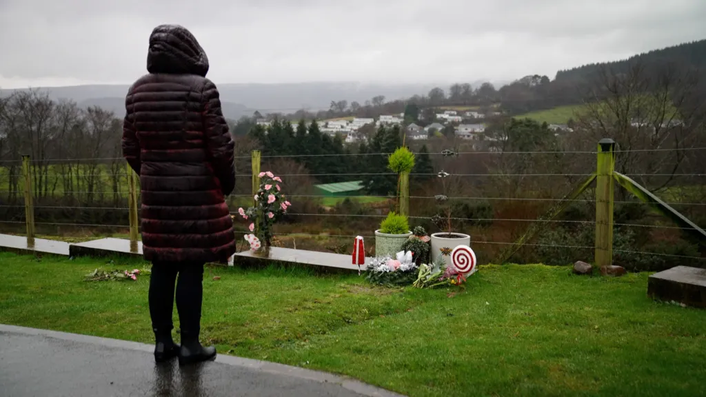 A woman in a burgundy jacket, with her hood up, looking at a grave on the edge of a cemetery. In the background is a view of trees, fields and houses.