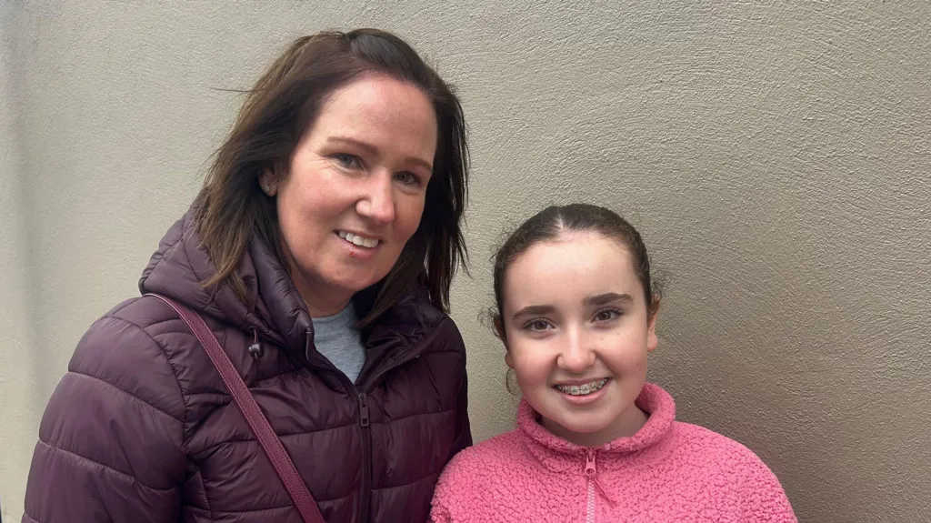 Pamela and Emily on the High Street, Pamela is wearing a purple coat, she has long brown hair. Emily is wearing a pink zip-up jumper and has tied up brown hair, she also has braces. 
