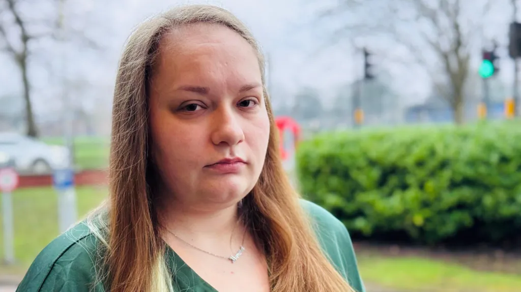 Georgia Spargo is outside, the background of trees and a road is out of focus.
She has long brown hair, is wearing a green dress and a silver necklace. She is looking straight at the camera, and is not smiling.