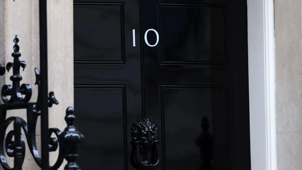 A black door in Downing Street with the Number 10 on it. A black railing can be seen in the foreground.