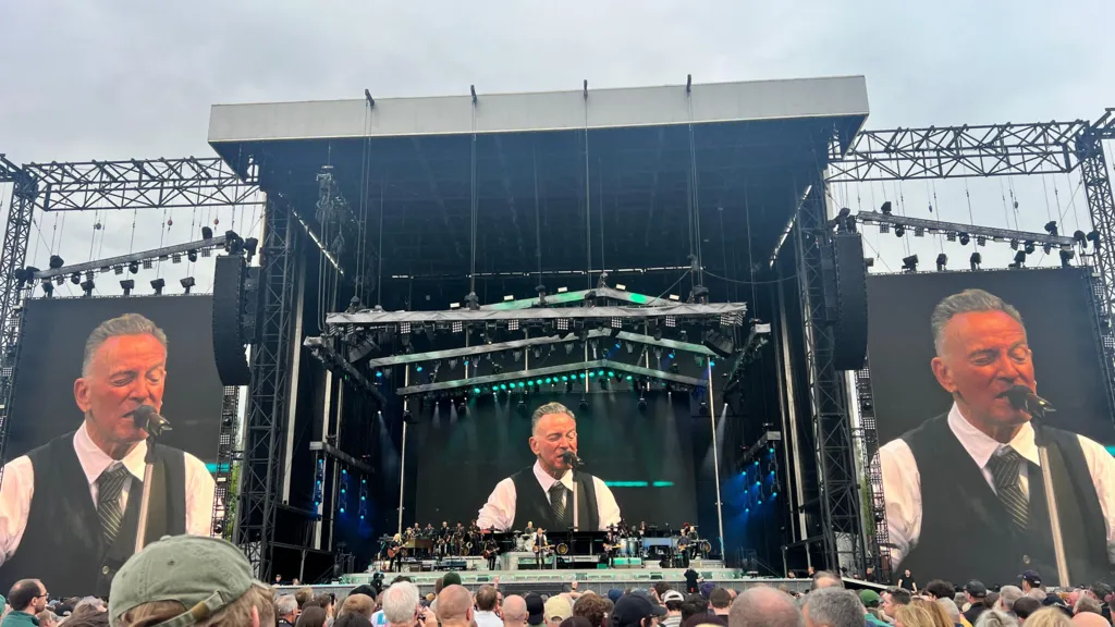 A picture taken from the crowd of three screens showing Bruce Springsteen playing on stage. Springsteen has short grey hair and is singing into a mic, he is wearing a white shirt, black tie and waist coat.