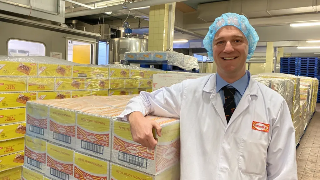 Stuart Louden wears a white protective coat and a food-hygiene hat. He's leaning on a pallet loaded with boxes of Tunnock's wafer bars.