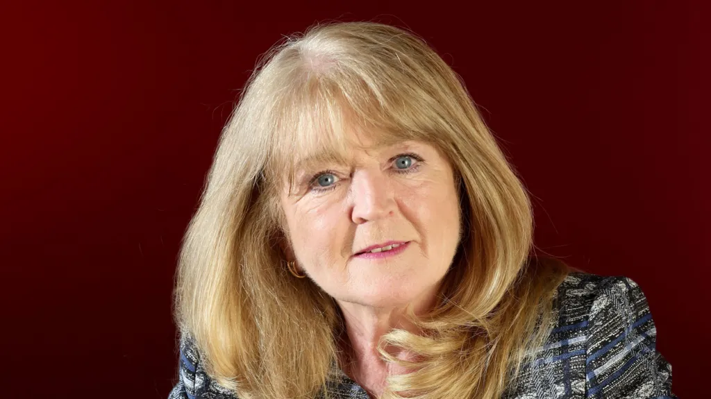 Geraldine McGahey. She has long blonde hair with a fringe, blue eyes, wearing a dark grey blazer. Behind her is a dark red background.