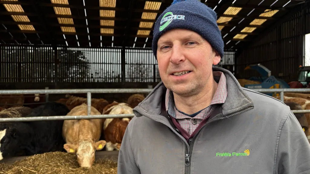 Fergal Watson, a man with short hair, looks to the camera on his farm in Cloughey, County Down. He is wearing a blue beanie hat with a green logo, a grey zipped jacket and a red checked shirt. He is standing in front of a large cattle shed.  Cattle are eating silage from a feeding trough. 
