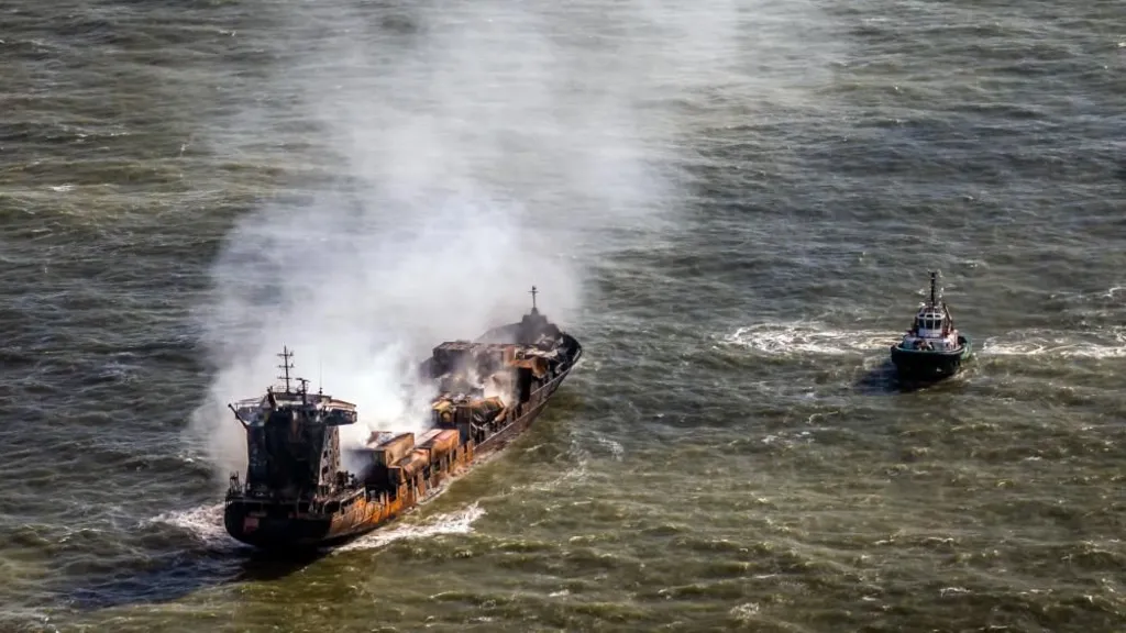 Smoke billows from a rust-coloured tanker ship floating in the sea. A tugboat floats nearby.