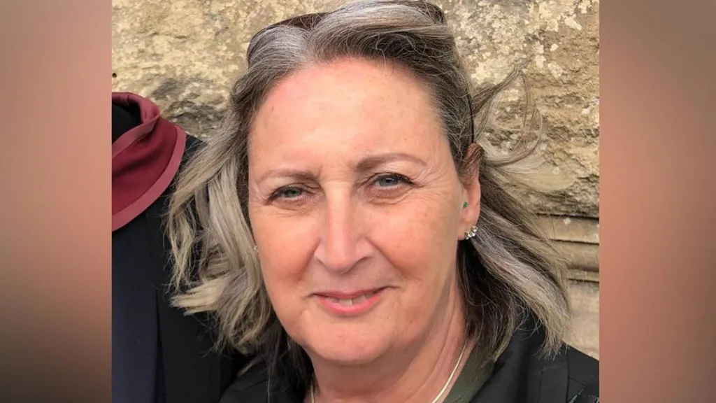 A woman with short grey hair wearing a black jacket, khaki green shirt, and a bag with a gold chain. She is smiling at the camera and has a pair of sunglasses on her head.