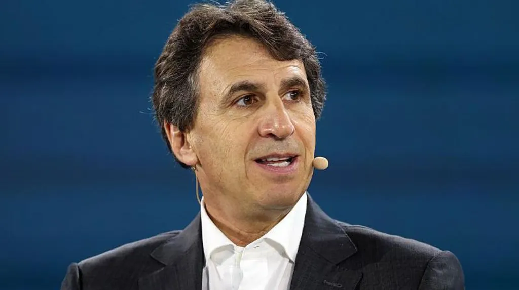 An image of businessman Marc Rowan looking to the right with a microphone attached to his face, while dressed in a black suit and sat in front of a blue backdrop.