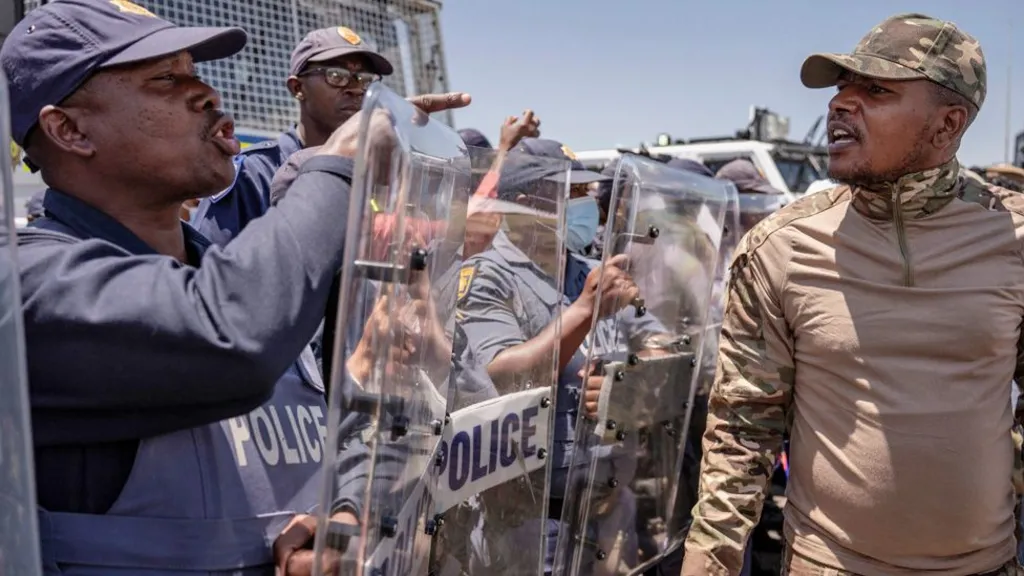 Riot police with shields confront an angry man from Operation Dudula in a camouflage top and cap. One of the officers is pointing a finger at him.