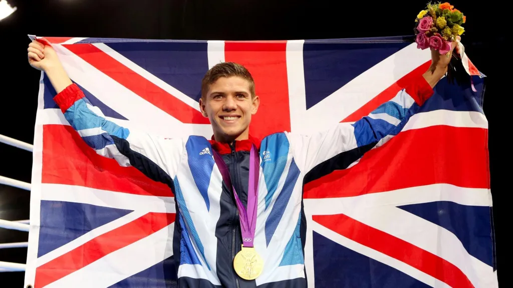 Luke Campbell holds a Union Flag aloft as he smiles at the camera. He is wearing a Team GB tracksuit and has a gold medal around his neck.