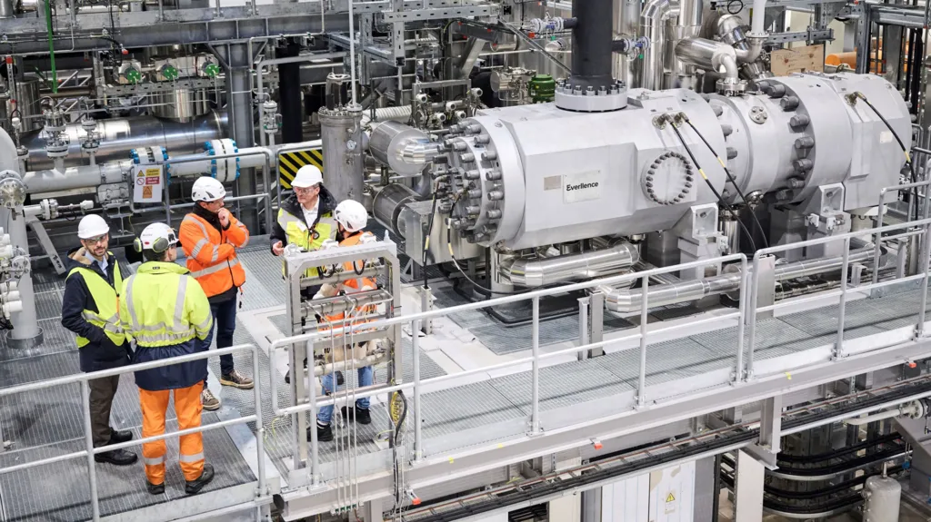 Five men in hi-viz jackets talk while standing next to heat pump machinery.