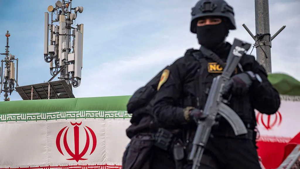 An armed Special Units police officer in Iran, dressed in black and holding a weapon, is standing on an armored vehicle while monitoring the surroundings. In the background, telecommunications towers and the Iranian flag can be seen. The photo was taken during a pro-government gathering in central Tehran.