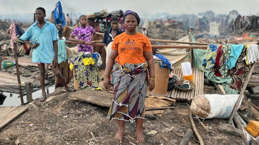 Anna Sobie, wearing an orange T-shirt and African print fabric wrapped around her waist, stands in the ruins of her house. Her children can be seen behind her and some clothes hanging on pieces of wood.