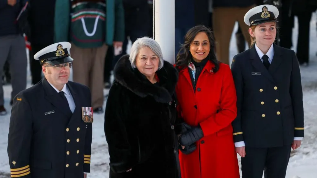 An image showing Foreign Minister Anita Anand and Governor General Mary Simon, with Canadian officers at either of their sides. Anand is wearing a bright red coat and black gloves, and she has shoulder-length wavy black hair. Simon is wearing a large fur black coat, and has a short white bob. 