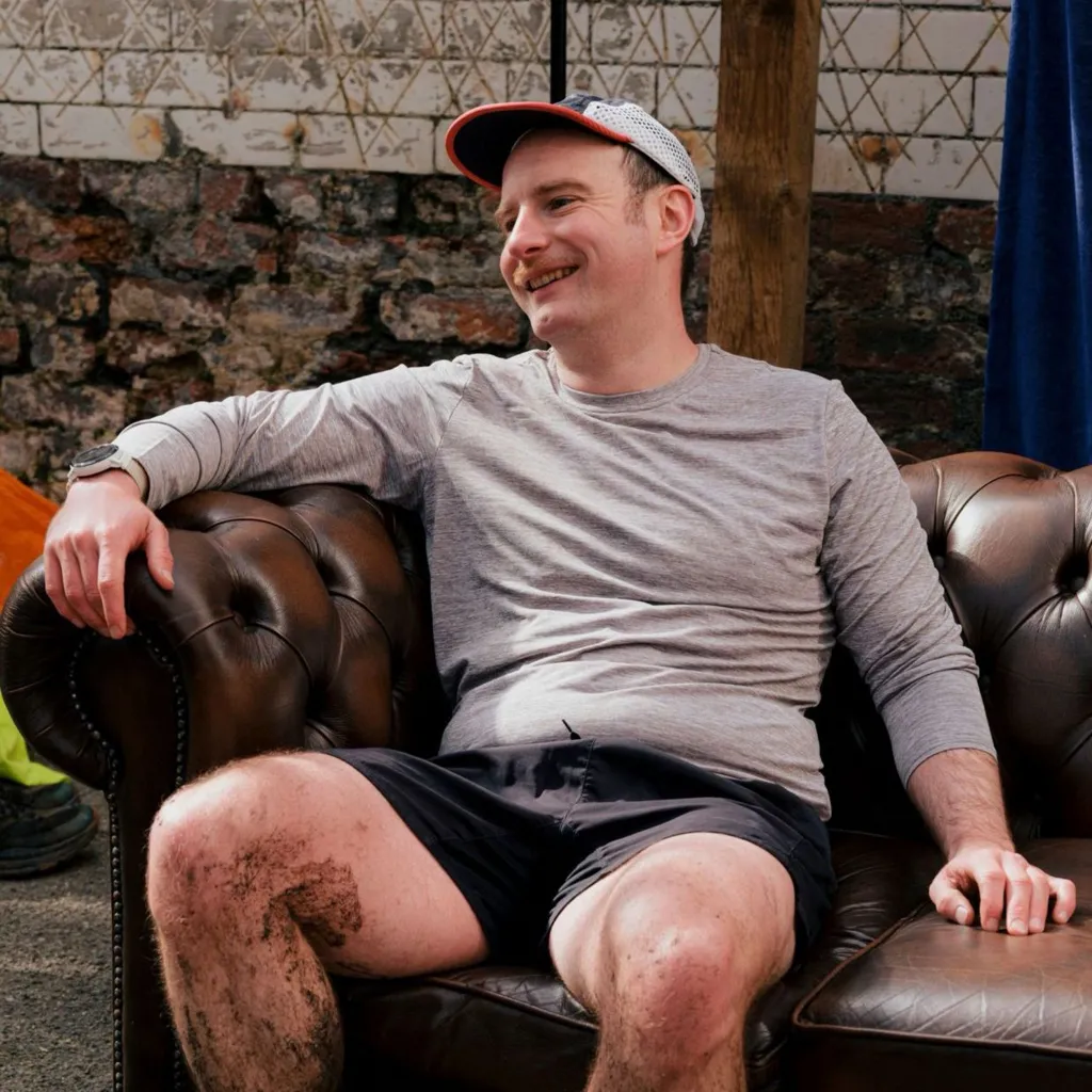 Xander, a 37 year old man is sat on a on a sofa in shorts and a running top. He's laughing and he's got a muddy leg