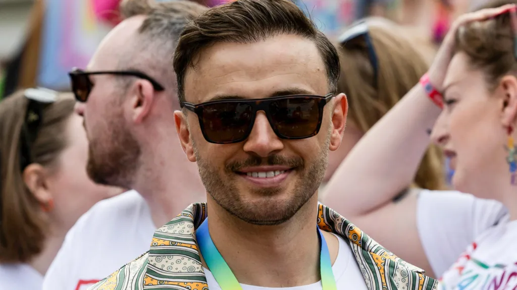 Zander Murray stands in a crowd at Edinburgh Pride 2023. He has dark hair and a beard and is wearing sunglasses and a rainbow lanyard around his neck. He is also wearing a green and yellow patterned shirt.