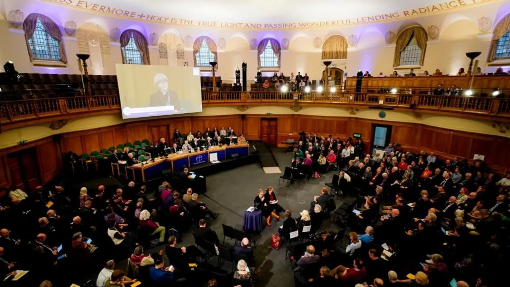 The Archbishop of Canterbury addresses the General Synod. She stands behind a long table with her image on a large screen above her. There are a lot of people in attendance.  