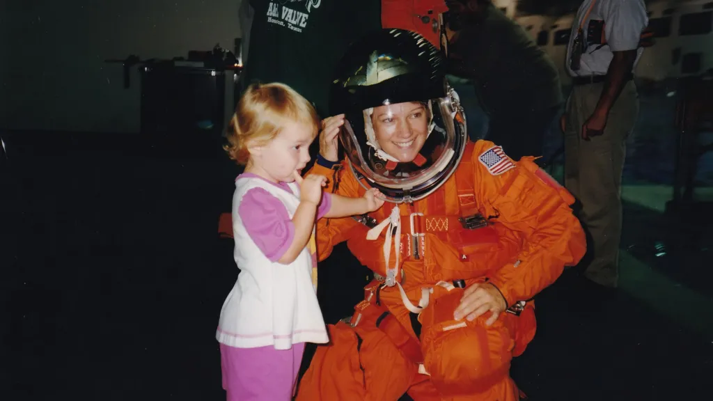 Astronaut Eileen Collins wears an orange spacesuit with a clear helmet. She smiles as she crouches next to her daughter, who is three years old. She is wearing a pink and white outfit and has her finger on her mouth and the other hand on her Mum's pace helmet. She looks shy.  
