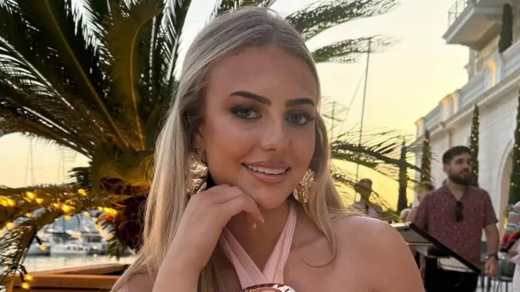 Daisy is sitting down with a palm tree in the background. Daisy has blonde hair- worn half up half down. She has a pink halter neck top and big gold earrings.