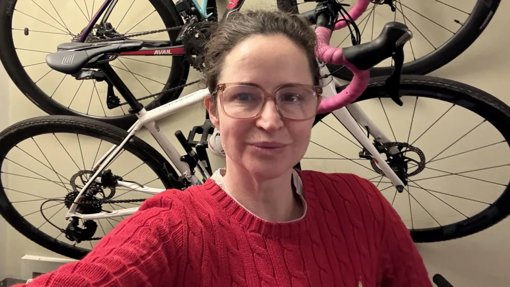 Wearing a red jumper and glasses, Kate Corden, stands in front of bicycles hanging on a wall.