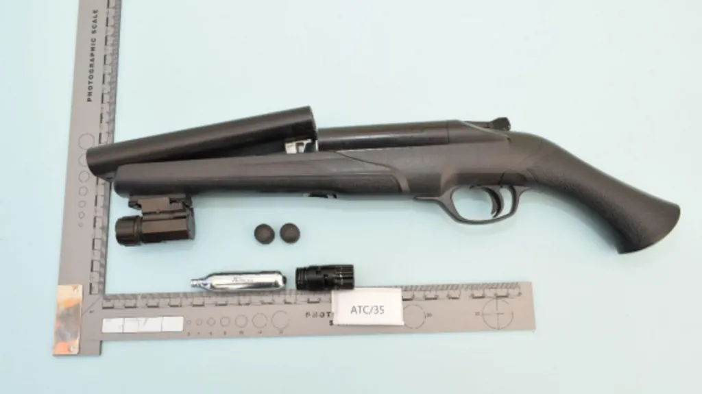 A collection of items laid out on a light blue surface and framed by a photographic measurement scale. The central object is a compact, black, break‑action firearm with its barrel open. The grip and frame are made of a dark synthetic material, and the trigger and trigger guard are clearly visible.
Below the firearm are a small metallic gas canister, two round black pellets or projectiles, and a black cylindrical attachment. Another larger black attachment is placed to the left of the firearm, positioned near the hinge of the barrel.
A labelled evidence marker reading “ATC/35” is placed alongside the items.