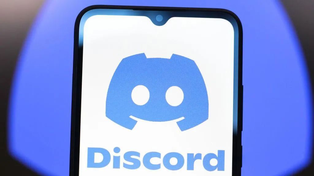Discord logo shown on a smartphone screen in front of a larger, blurred version of the company's blue logo.