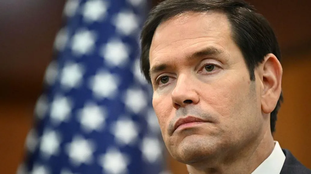 An image of the US Secretary of State, Marco Rubio, speaking into a microphone while wearing a navy suit with a red tie.