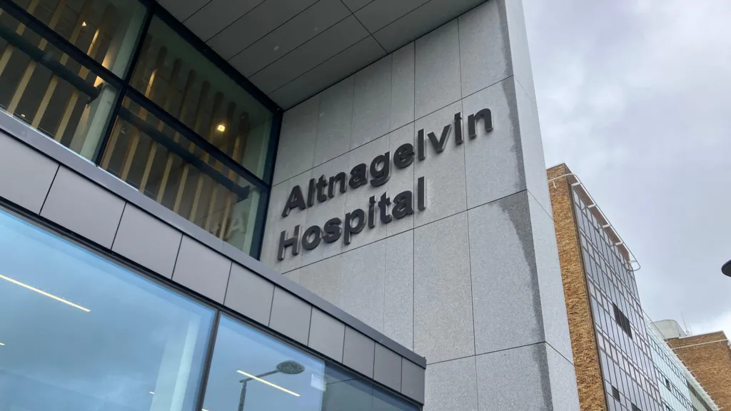 A close up of a sign outside Altnagelvin Hospital, which says the name of the building in large lettering. It is a tall building with a mix of facades including brown brick and grey cladding.