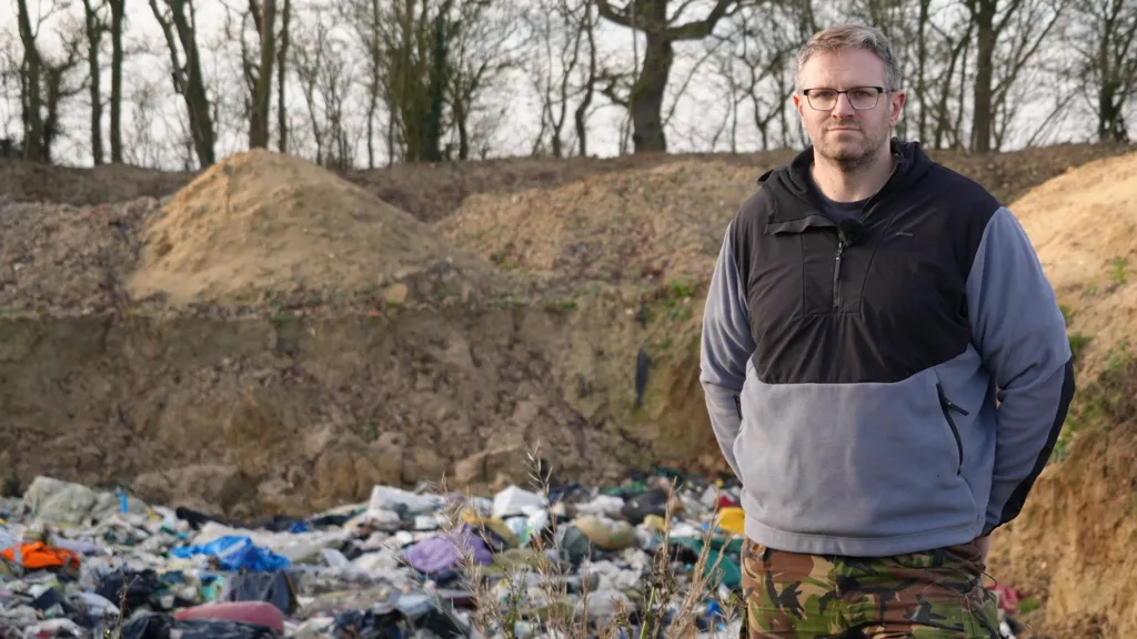 Peter Dive stands beside a pit filled with waste. He is wearing a black and grey fleece top and camouflage trousers. He has his hands behind his back. He is looking directly at the camera and has stubble and black-framed glasses.